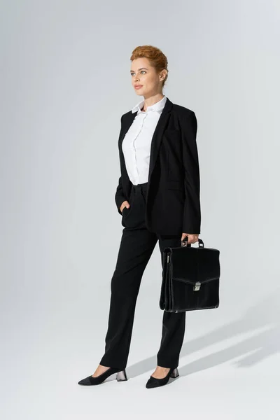 Повна довжина позитивної бізнес-леді в чорному костюмі позує з портфелем і рукою в кишені на сірому фоні — стокове фото