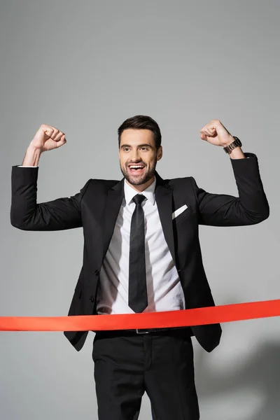 Overjoyed businessman in black blazer showing win gesture near finish ribbon on grey background — Stock Photo