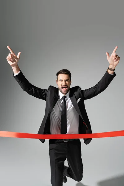 Успешный бизнесмен радуясь и показывая жест триумфа рядом с красной лентой финиша на сером фоне — стоковое фото