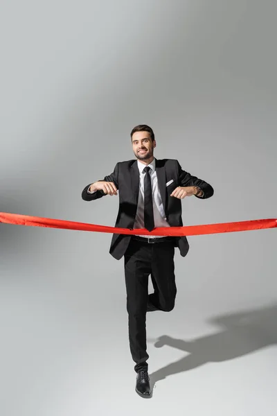 Повна довжина щасливого бізнесмена в чорному костюмі біжить і перетинає червону стрічку на сірому фоні — стокове фото