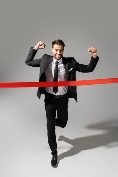 Full length of overjoyed businessman crossing finish ribbon while running and showing win gesture on grey background — Stock Photo
