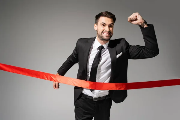 Успішний і схвильований бізнесмен, що показує жест перемоги і перетинає червону фінішну стрічку ізольовано на сірому — стокове фото