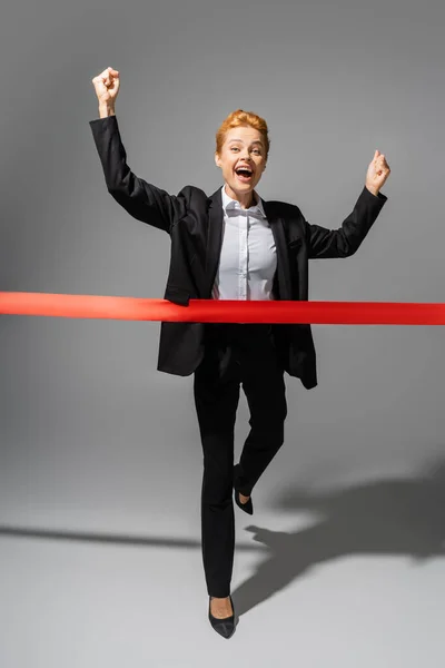 Volle Länge der fröhlichen Geschäftsfrau jubeln und überqueren Finish Band auf grauem Hintergrund — Stockfoto