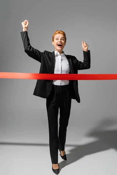 Full length of overjoyed businesswoman in black suit crossing finish ribbon on grey background — Stock Photo