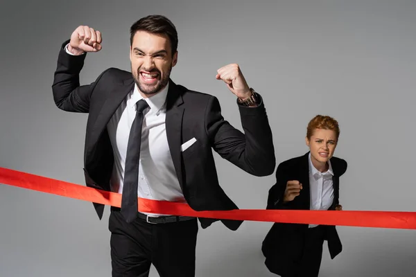 Overjoyed businessman in black suit showing win gesture near sad woman running on grey background — Stock Photo