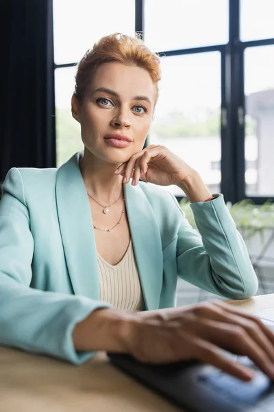 Stylish redhead businesswoman sitting near blurred laptop and looking at camera in office — Stock Photo