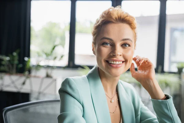 Portrait of cheerful redhead businesswoman in stylish blazer looking at camera in office — Stock Photo