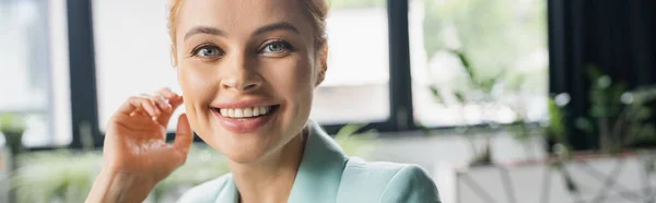 Portrait of cheerful businesswoman holding hand near face and smiling at camera in office, banner — Stock Photo