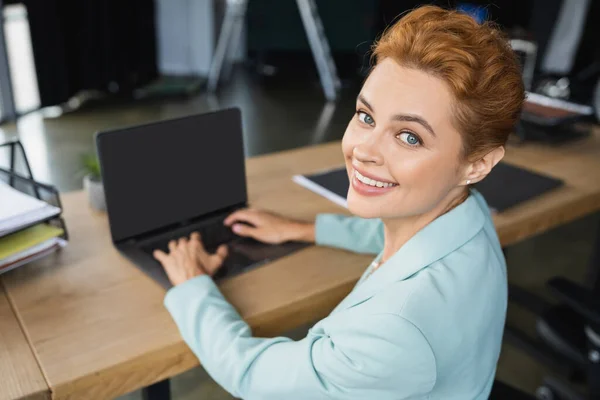Alegre pelirroja mujer de negocios escribiendo en borrosa portátil con pantalla en blanco y sonriendo a la cámara en la oficina - foto de stock