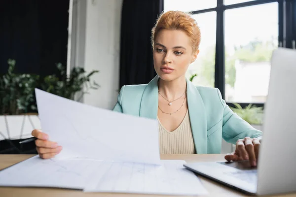 Fokussierte rothaarige Geschäftsfrau arbeitet mit verschwommenen Papieren und Laptop im Büro — Stockfoto