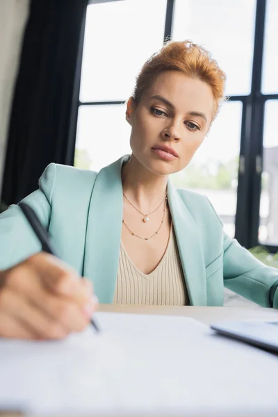Concentrada pelirroja mujer de negocios en elegante blazer escritura en la oficina en primer plano borrosa - foto de stock