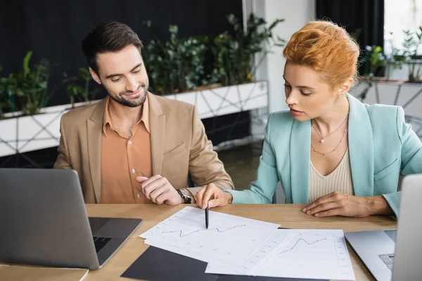 Bearded businessman smiling near laptops and redhead colleague working with charts in office — Stock Photo