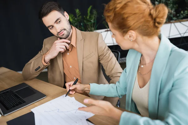 Вдумчивый менеджер смотрит на рыжую деловую женщину, указывающую на графики на размытом переднем плане в офисе — стоковое фото