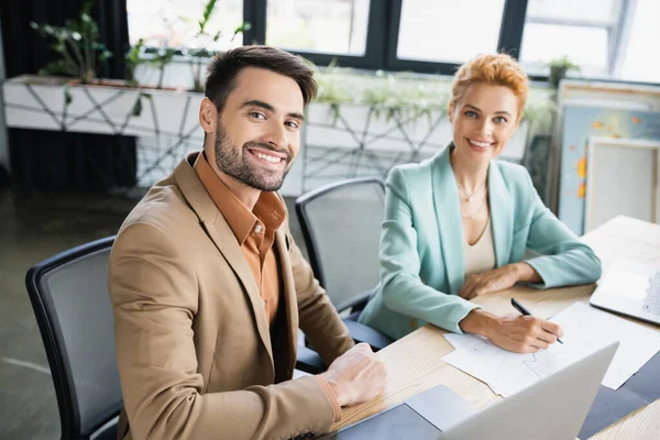 Happy and stylish business partners looking at camera near documents and laptops in office — Stock Photo