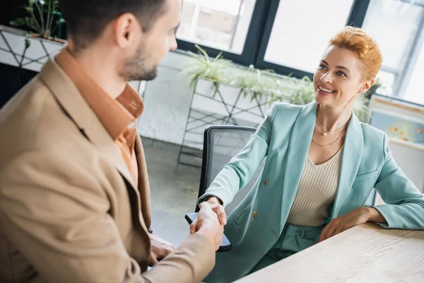 Joyful redhead businesswoman shaking hands with blurred partner in office — Stock Photo