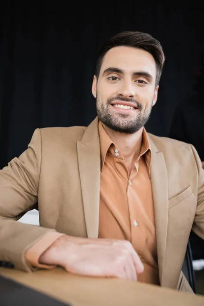 Bearded businessman in beige blazer smiling at camera while sitting in office — Stock Photo