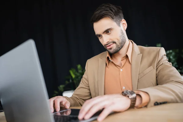 Homme d'affaires souriant en blazer beige travaillant sur ordinateur portable flou dans le bureau — Photo de stock