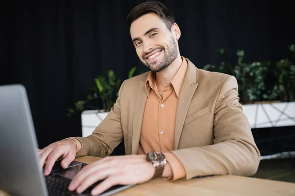 Веселий бородатий бізнесмен друкує на ноутбуці і дивиться на камеру на робочому місці в офісі — стокове фото
