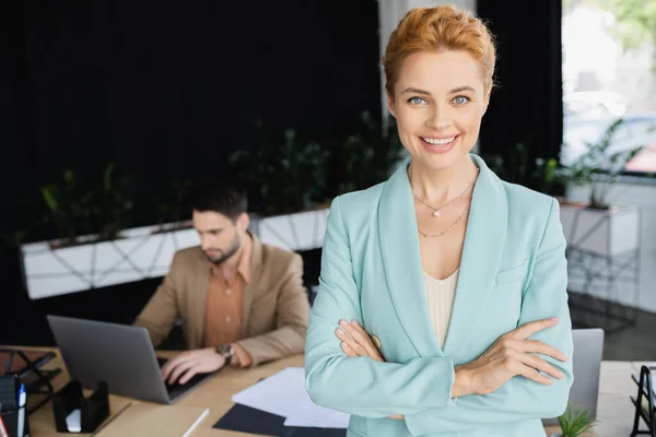 Довольная деловая женщина в модном блейзере, смотрящая в камеру рядом с коллегой, работающим на ноутбуке в офисе — стоковое фото