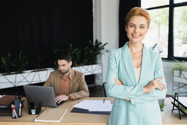 Руда бізнес-леді зі складеними руками посміхається на камеру біля колеги, що працює на ноутбуці в офісі — стокове фото
