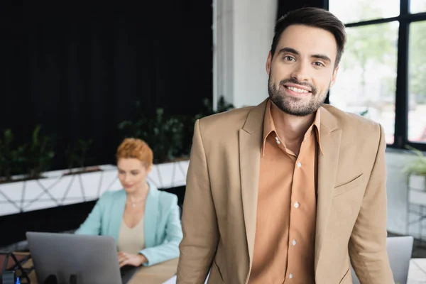 Homme d'affaires barbu dans blazer élégant souriant à la caméra près de collègue flou travaillant sur ordinateur portable dans le bureau — Photo de stock