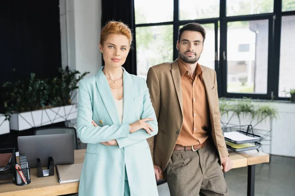 Positive business colleagues standing at work desk in modern office and looking at camera — Stock Photo