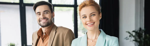 Portrait of bearded man and redhead woman in blazers smiling at camera in office, banner — Stock Photo