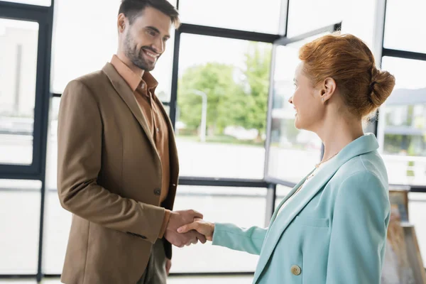Pleased businessman in trendy suit shaking hands with redhead manager in office — Stock Photo