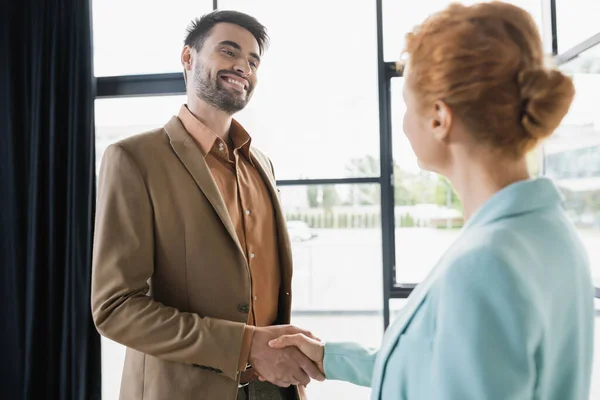 Успешный бизнесмен улыбается и пожимает руку рыжей женщине в офисе — стоковое фото