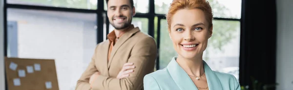 Успешная рыжая деловая женщина смотрит в камеру рядом с коллегой улыбается на размытом фоне в офисе, баннер — стоковое фото