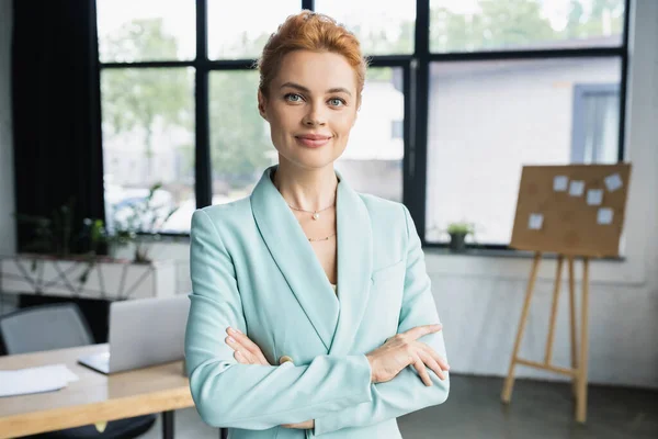 Glückliche rothaarige Geschäftsfrau in stylischem Blazer, die Arme verschränkt und im Büro in die Kamera blickt — Stockfoto