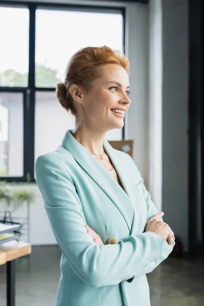 Exitosa mujer de negocios de pie con los brazos cruzados mientras sonríe y mira hacia otro lado en la oficina - foto de stock