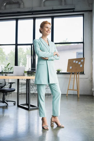 Longitud completa de alegre pelirroja mujer de negocios en elegantes pantalones de pie con los brazos cruzados en la oficina moderna - foto de stock