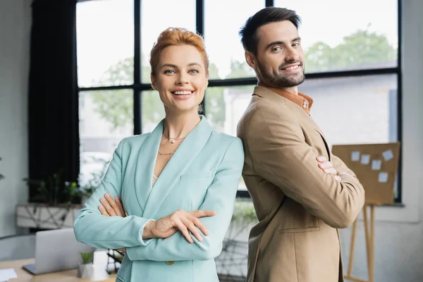 Радостные деловые партнеры, улыбающиеся в камеру, позируя со сложенными руками в офисе — стоковое фото