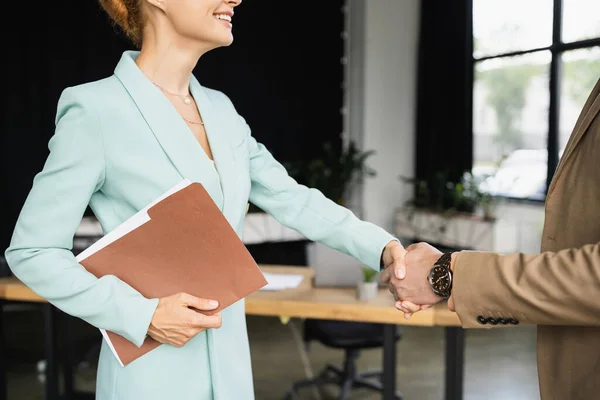 Visão cortada de mulher de negócios feliz com pasta apertando as mãos com o gerente no escritório — Fotografia de Stock