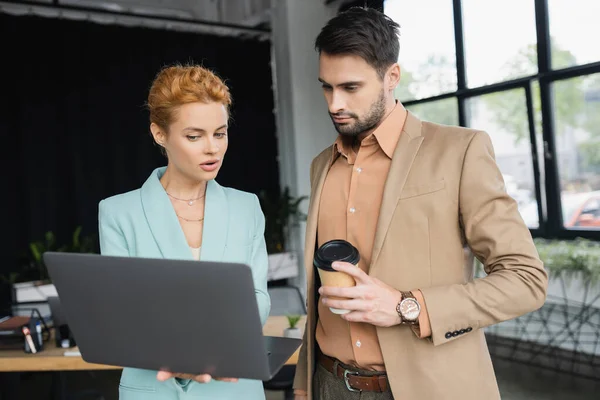 Redhead businesswoman in eyeglasses showing laptop to colleague with paper cup in office — Stock Photo