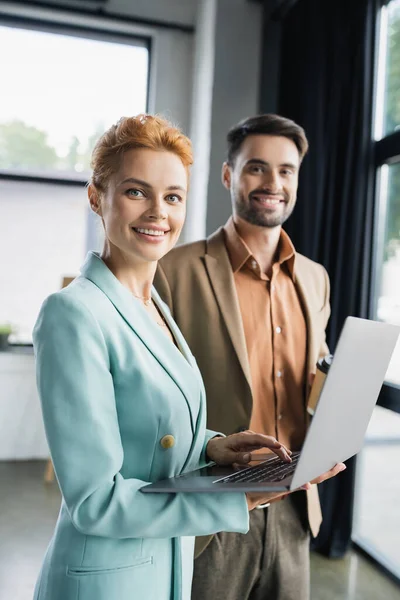 Freudige Geschäftspartner in trendigen Blazern lächeln in die Kamera neben dem Laptop im Büro — Stockfoto