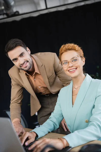 Happy and stylish business people looking at camera near blurred laptop in office — Stock Photo