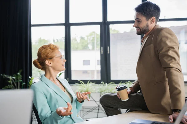 Счастливая деловая женщина разговаривает с коллегой сидя на рабочем столе с бумажной чашкой в офисе — стоковое фото