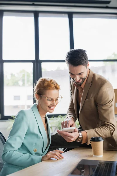 Smiling bearded businessman showing smartphone to woman in eyeglasses in office — Stock Photo