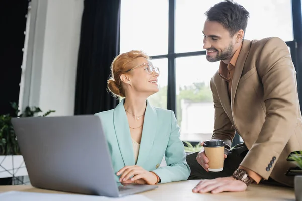 Happy businesswoman in eyeglasses working on laptop and looking at bearded colleague with coffee to go in office — Stock Photo