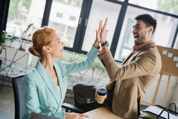 Cheerful man with coffee to go giving high five to businesswoman sitting at workplace in office — Stock Photo