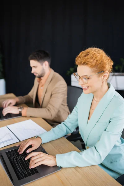 Руда бізнес-леді в окулярах, друкуючи на ноутбуці біля розмитого колеги в офісі — стокове фото