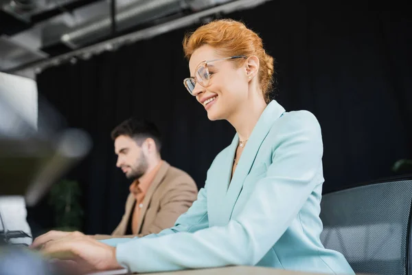 Счастливая рыжая деловая женщина в очках, работающая рядом с коллегой на размытом фоне в офисе — стоковое фото