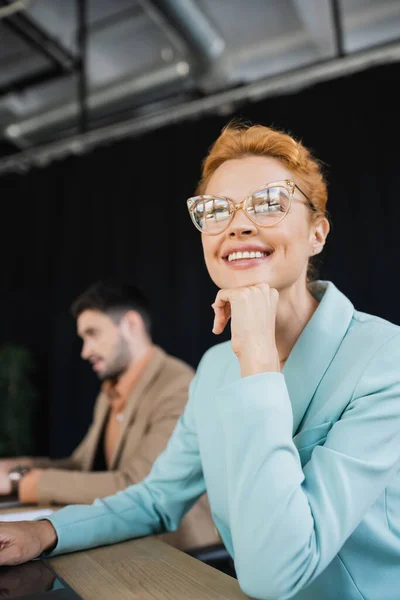 Feliz y soñadora mujer de negocios en gafas mirando hacia otro lado cerca borrosa colega que trabaja en la oficina - foto de stock