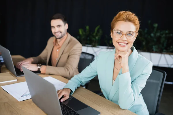 Joyful businesswoman smiling at camera near laptop and blurred colleague in office — Stock Photo
