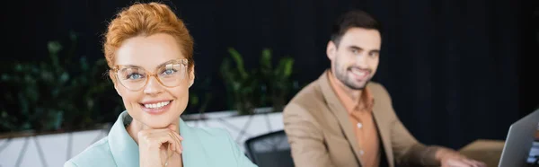 Portrait of pleased businesswoman in eyeglasses looking at camera near blurred colleague in office, banner — Stock Photo