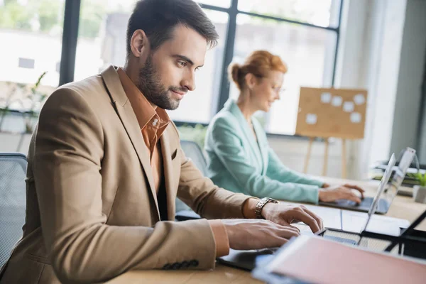 Позитивний бородатий бізнесмен друкує на ноутбуці біля розмитого колеги в офісі — стокове фото
