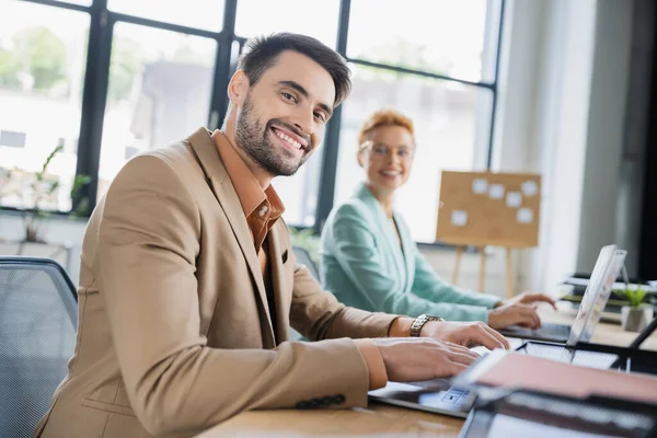 Cheerful bearded manager looking at camera while working on laptop near blurred businesswoman in office — Stock Photo