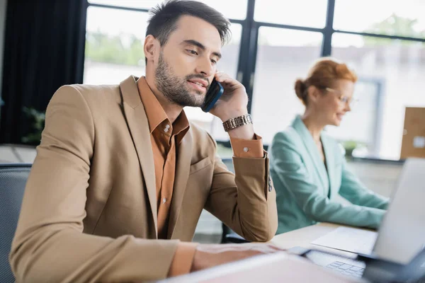 Бородатий бізнесмен говорить на смартфоні біля ноутбука та жінки на розмитому фоні в офісі — стокове фото